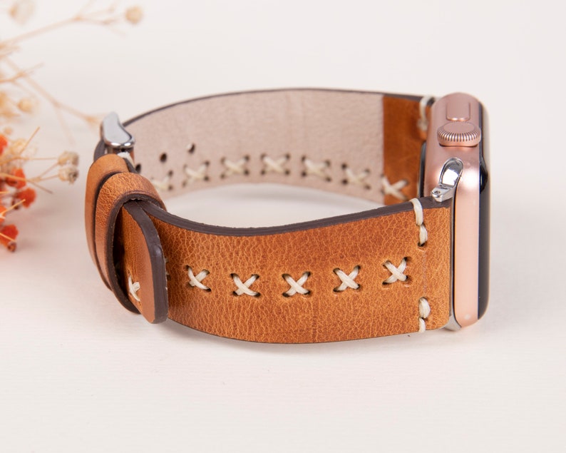 May include: A brown leather watch band with white stitching. The band has a stitched pattern of X's. The band is attached to a rose gold Apple Watch.