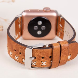 May include: Tan leather Apple Watch band with white stitching and a silver buckle. The band has a cross-stitch pattern.