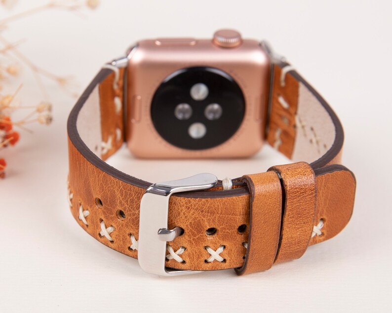 May include: Tan leather Apple Watch band with white stitching and a silver buckle. The band has a cross-stitch pattern.