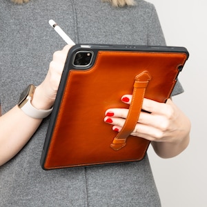 Puede incluir: Una funda de tableta de cuero marrón con un marco negro y una correa de mano. La funda la sostiene una persona que lleva un vestido gris y un reloj dorado con una correa blanca. También se ve un lápiz óptico blanco.