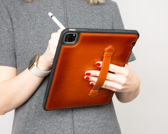 May include: A brown leather tablet case with a black frame and a hand strap. The case is being held by a person wearing a grey dress and a gold watch with a white strap. A white stylus is also visible.
