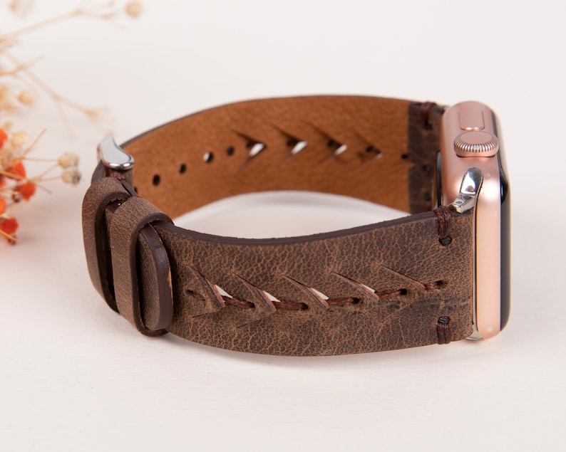 Puede incluir: Una correa de reloj de cuero marr&oacute;n con un dise&ntilde;o trenzado y un Apple Watch de oro rosa.