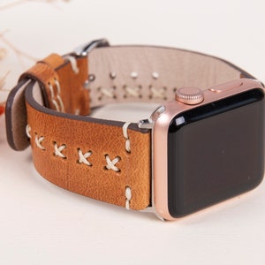 May include: A brown leather Apple Watch band with white stitching. The band has a rose gold Apple Watch attached to it.