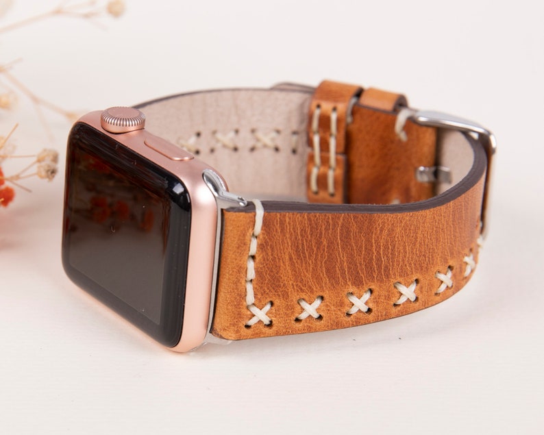 May include: A brown leather Apple Watch band with white stitching. The band has a classic look and is compatible with Apple Watch models.
