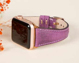 May include: A rose gold Apple Watch with a purple leather band. The band has white stitching and a rose gold buckle.
