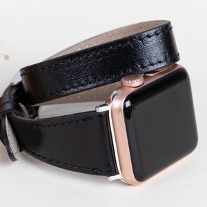 Puede incluir: Correa de reloj de cuero negro de doble envoltura combinada con un reloj inteligente de oro rosa. La correa está cosida y tiene un cierre plateado. La esfera del reloj es negra.