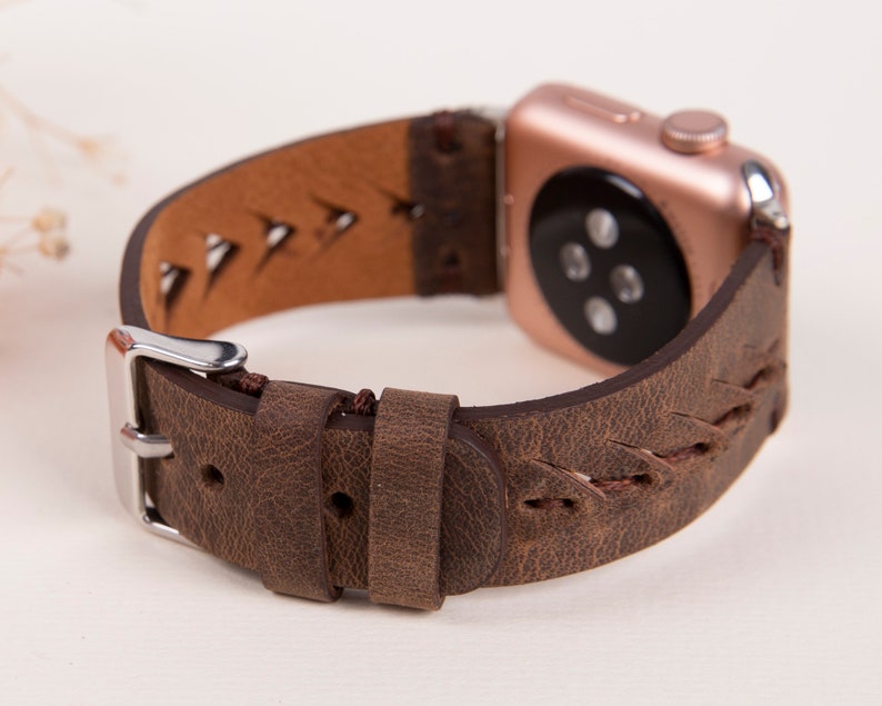 Puede incluir: Una correa de reloj de cuero marr&oacute;n con un dise&ntilde;o trenzado y una hebilla plateada. La correa est&aacute; dise&ntilde;ada para un Apple Watch y tiene una esfera de reloj de oro rosa.