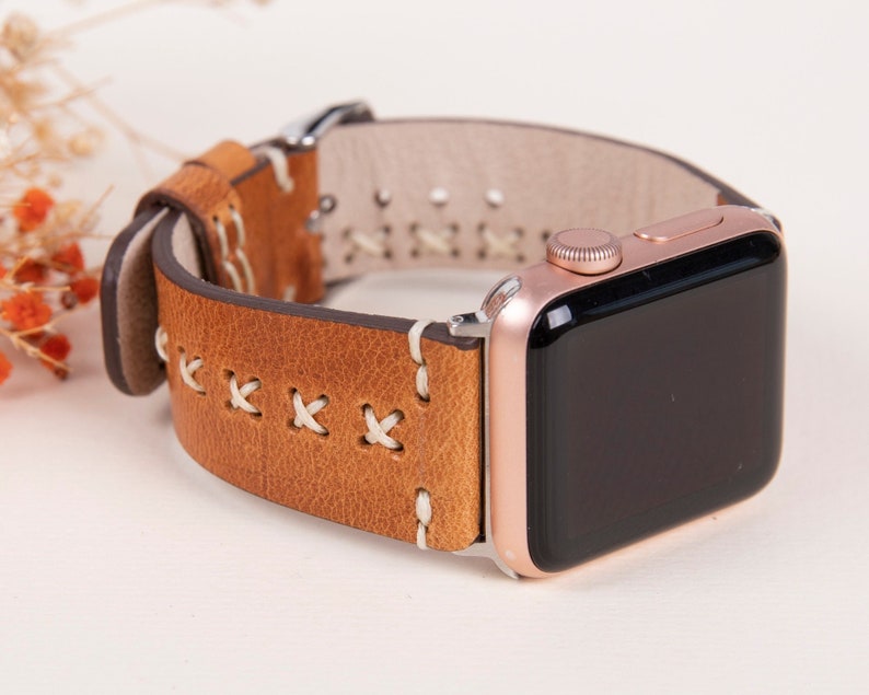 May include: A brown leather watch band with white stitching for an Apple Watch. The watch band has a rose gold Apple Watch attached to it.