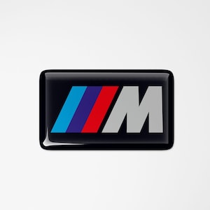 Könnte beinhalten: Ein glänzend schwarzes rechteckiges Emblem mit den Buchstaben "M" in Grau und blauen, roten und weißen Streifen. Das Emblem befindet sich vor einem weißen Hintergrund.