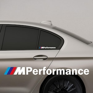 Op de afbeelding: Een zilveren auto met een zwarte band en raam. De auto heeft een witte "MPerformance" sticker met blauwe, rode en witte strepen. Een kleinere sticker staat op het raam.