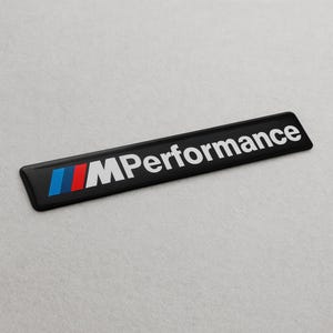Könnte beinhalten: Ein schwarzes rechteckiges Emblem mit dem weißen Text "M Performance". Das Emblem hat blaue, rote und hellblaue Streifen auf der linken Seite. Wahrscheinlich ein Autoabzeichen.