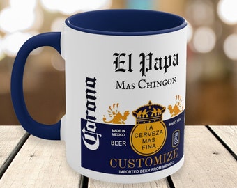 May include: White ceramic mug with a navy blue interior and handle. The mug features the text "El Papa Mas Chingon" and "Corona" branding, along with other text and graphics. The mug is designed to hold 20 fl oz of liquid.