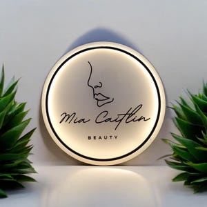 Puede incluir: Un letrero redondo e iluminado con un borde negro. El letrero presenta un dibujo lineal minimalista de un rostro y las palabras "Mia Caitlin BEAUTY" en escritura cursiva. Plantas verdes enmarcan el letrero.