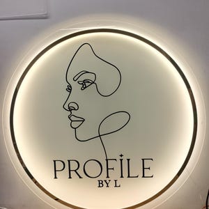 Può includere: Un cartello circolare bianco con un bordo dorato e un disegno a linea nera di un viso di donna. Il testo "PROFILE BY L" è stampato in nero sotto il disegno.