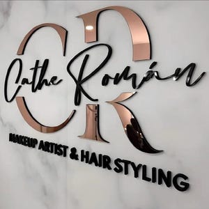 Puede incluir: Un letrero negro y dorado rosa con el texto "Cathe Román" y "Makeup Artist & Hair Styling".