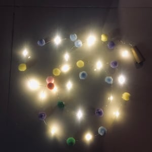 Puede incluir: Una cadena de luces de hadas con pompones de colores. Las luces son blancas y los pompones son en tonos de azul, verde, amarillo y rosa.