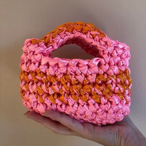 May include: A small pink and orange crocheted handbag with a woven handle.