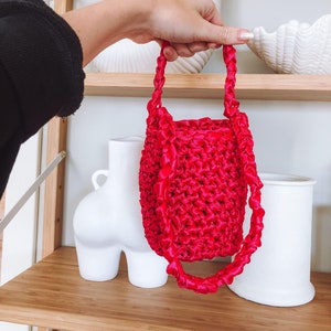 Może przedstawiać: Jasno różowa torba na szydełku z grubym, plecionym paskiem. Torba wykonana jest z miękkiego, fakturowanego materiału i ma cylindryczny kształt.