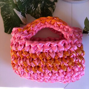 May include: A small pink and orange crocheted bag with a handle. The bag is made of woven ribbon.
