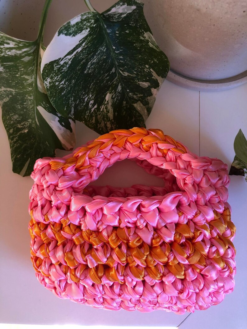 May include: A small pink and orange crocheted bag with a handle. The bag is made of woven ribbon.