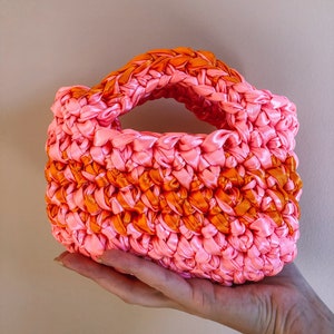 Könnte beinhalten: Eine kleine gehäkelte Handtasche in Pink und Orange mit einem einzigen Henkel. Die Tasche ist aus dickem, gewebtem Band gefertigt.