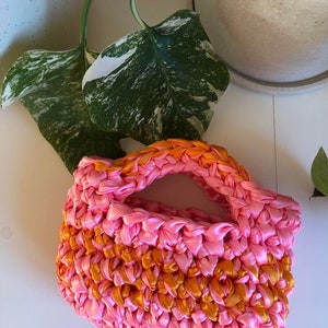 May include: A small pink and orange crocheted bag with a handle. The bag is made from woven ribbon.
