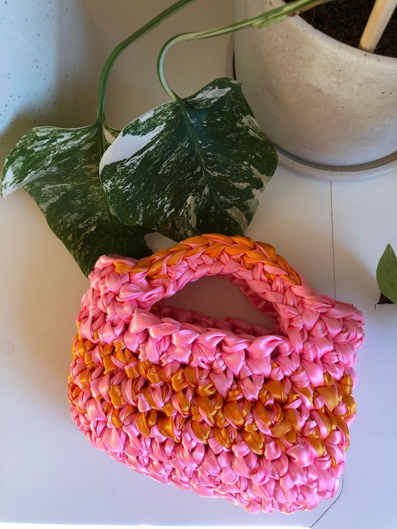 May include: A small pink and orange crocheted bag with a handle. The bag is made from woven ribbon.