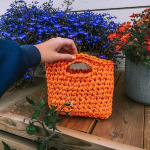 May include: A vibrant orange crocheted handbag, square in shape, with a handle opening. The bag is made from a woven material. The background features blue and orange flowers and a wooden surface.