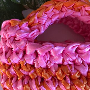 May include: A pink and orange crocheted bag made with satin ribbon. The bag has a woven texture and is made with a thick, chunky yarn.