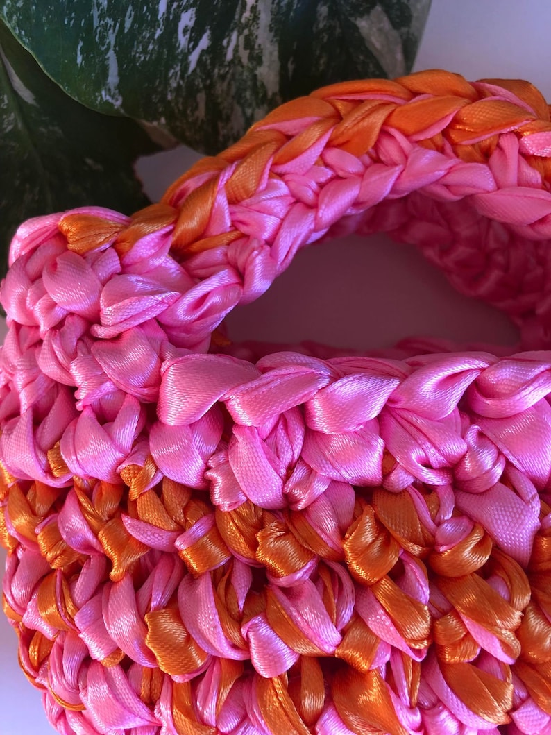 May include: A pink and orange crocheted bag made with satin ribbon. The bag has a woven texture and is made with a thick, chunky yarn.