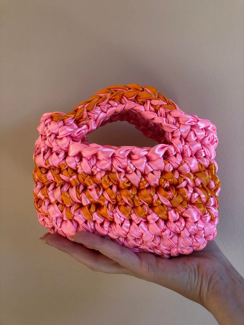 May include: A small pink and orange crocheted handbag with a woven handle.