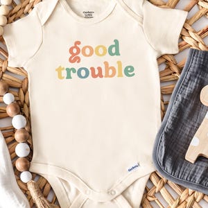 May include: Cream-colored baby onesie with the words "good trouble" in colorful retro font. The onesie is next to a gray muslin burp cloth and a wooden giraffe toy. A pacifier and wooden beads are also in the image.