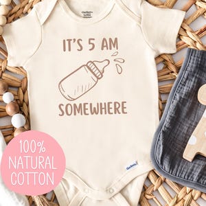 May include: A white baby onesie with the text "It's 5 AM Somewhere" and a drawing of a baby bottle. The onesie is made of 100% natural cotton.