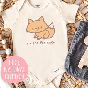 May include: Cream-colored baby onesie with a cartoon fox illustration and the phrase "oh, for fox sake." A gray muslin swaddle and a wooden giraffe toy are next to the onesie. The onesie is made of 100% natural cotton.