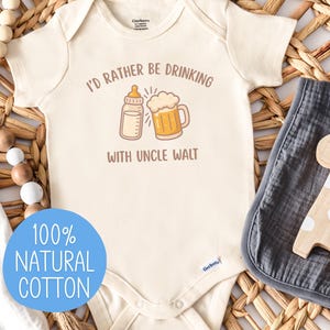May include: Cream-colored baby onesie with the phrase "I'd rather be drinking with Uncle Walt" and a graphic of a baby bottle and beer mug. The onesie is made of 100% natural cotton and is displayed with a gray burp cloth and a wooden giraffe toy.