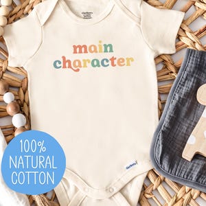 May include: A white baby onesie with the text "main character" in rainbow colors. The onesie is made of 100% natural cotton.