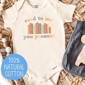 May include: Cream-colored baby onesie with the text "read to me you peasant" above a row of colorful books. The onesie is made of 100% natural cotton. A wooden giraffe toy and a gray muslin cloth are next to the onesie.