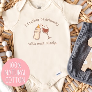 May include: Cream-colored baby onesie with the text "I'd rather be drinking with Aunt Mindy." A bottle and wine glass graphic are above the text. A wooden giraffe toy and a gray muslin cloth are next to the onesie. A pink circle with the text "100% NATURAL COTTON" is in the lower left.
