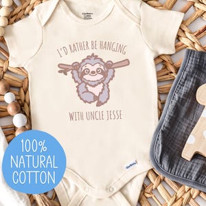May include: Cream-colored baby onesie with a sloth graphic and the text "I'd rather be hanging with Uncle Jesse." A blue circle reads "100% Natural Cotton." A wooden giraffe toy and a gray muslin cloth are also present.