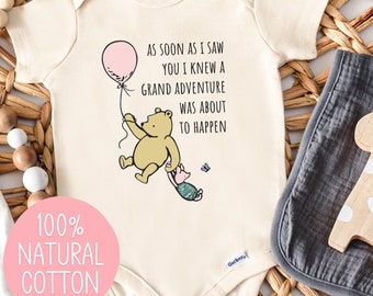 May include: A white baby onesie with a Winnie the Pooh illustration and the text "As soon as I saw you I knew a grand adventure was about to happen". The onesie is on a woven basket with a wooden giraffe toy and a gray burp cloth.