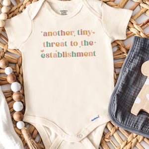 May include: A cream-colored short-sleeve baby bodysuit featuring the text "another tiny threat to the establishment" in multi-colored letters, accompanied by a wooden giraffe toy, a pacifier, and a string of wooden beads.