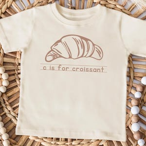 May include: Cream-colored toddler t-shirt with a brown illustration of a croissant and the text "c is for croissant". The shirt is displayed on a woven wicker surface, with decorative beads and a pink border.