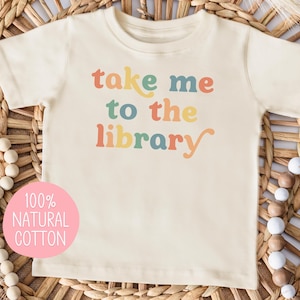 May include: Cream-colored t-shirt with the phrase "take me to the library" in colorful letters. A pink circle with the text "100% NATURAL COTTON" is on the bottom left. The shirt is displayed on a woven background with decorative beads.