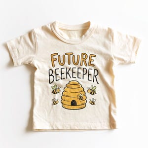 May include: A cream-colored short-sleeve t-shirt featuring the text "FUTURE BEEKEEPER" in black and gold lettering. Below the text is a graphic illustration of a yellow beehive surrounded by five small, smiling bees with green wings.