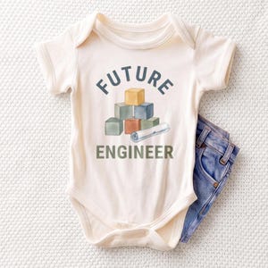 May include: A cream-colored short-sleeve baby bodysuit featuring a graphic of stacked colorful blocks and a rolled blueprint. The text FUTURE ENGINEER is printed in dark blue and green. A pair of blue denim jeans is tucked underneath the bodysuit.