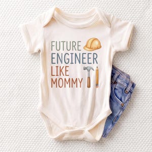 May include: A cream-colored baby bodysuit featuring the text FUTURE ENGINEER LIKE MOMMY in blue and brown lettering. The design includes a yellow hard hat, a grey hammer, and a brown pencil. A pair of blue denim jeans is tucked underneath the bodysuit.