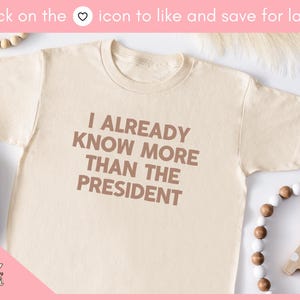 May include: A beige t-shirt with the text "I ALREADY KNOW MORE THAN THE PRESIDENT" printed in brown on the front.