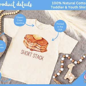 Op de afbeelding: Cr&egrave;mekleurig T-shirt voor peuters en jongeren met een ronde hals en korte mouwen. Het shirt heeft een aquarel illustratie van een kleine stapel pannenkoeken met boter en siroop, met de tekst "SHORT STACK". Gedrukt in de VS.