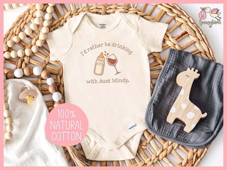 May include: Cream-colored baby onesie with the text "I'd rather be drinking with Aunt Mindy." A bottle and wine glass graphic are above the text. A wooden giraffe toy and a gray muslin cloth are next to the onesie. A pink circle with the text "100% NATURAL COTTON" is in the lower left.