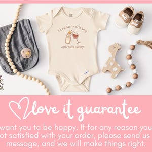 May include: A cream-colored baby onesie with the text "I'd rather be drinking with Aunt Becky." is displayed with baby shoes, a bib, a pacifier, and a wooden giraffe toy. A wooden bead garland and a pink background complete the scene.